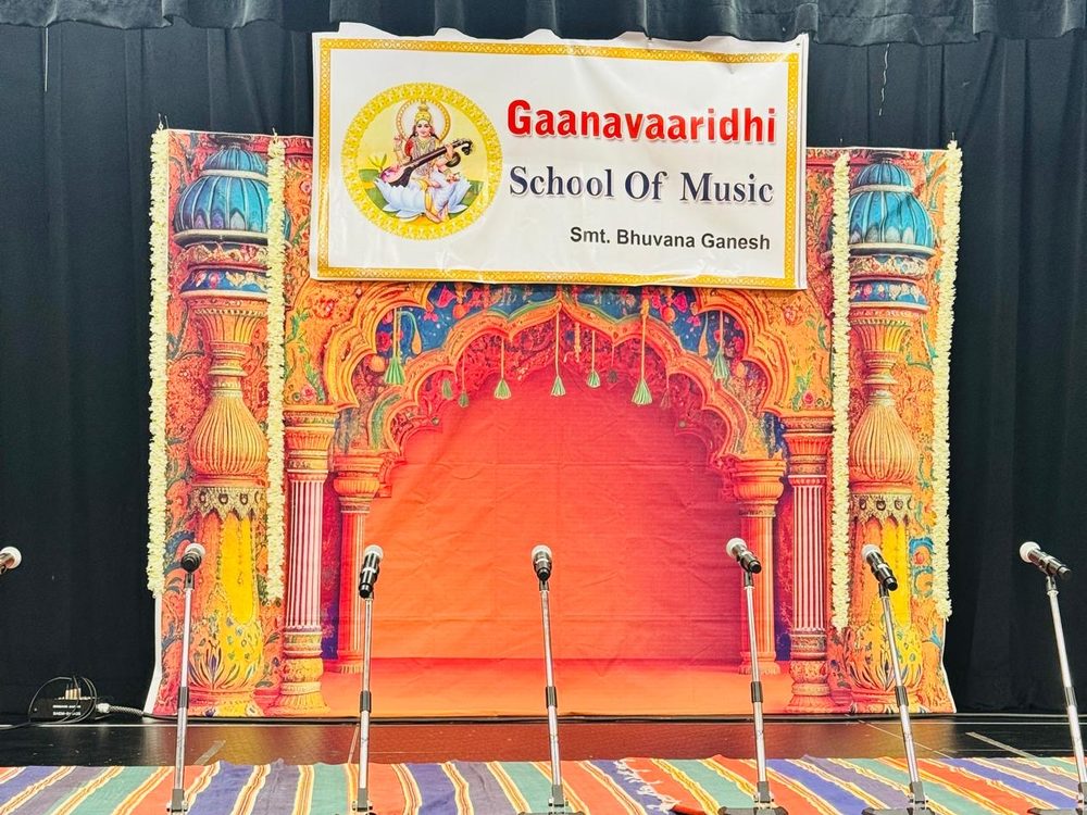 Gaanavaaridhi School of Music stage set for a concert: the school banner with Goddess Saraswati above a decorated temple-arch backdrop, jasmine garlands, and a row of microphones